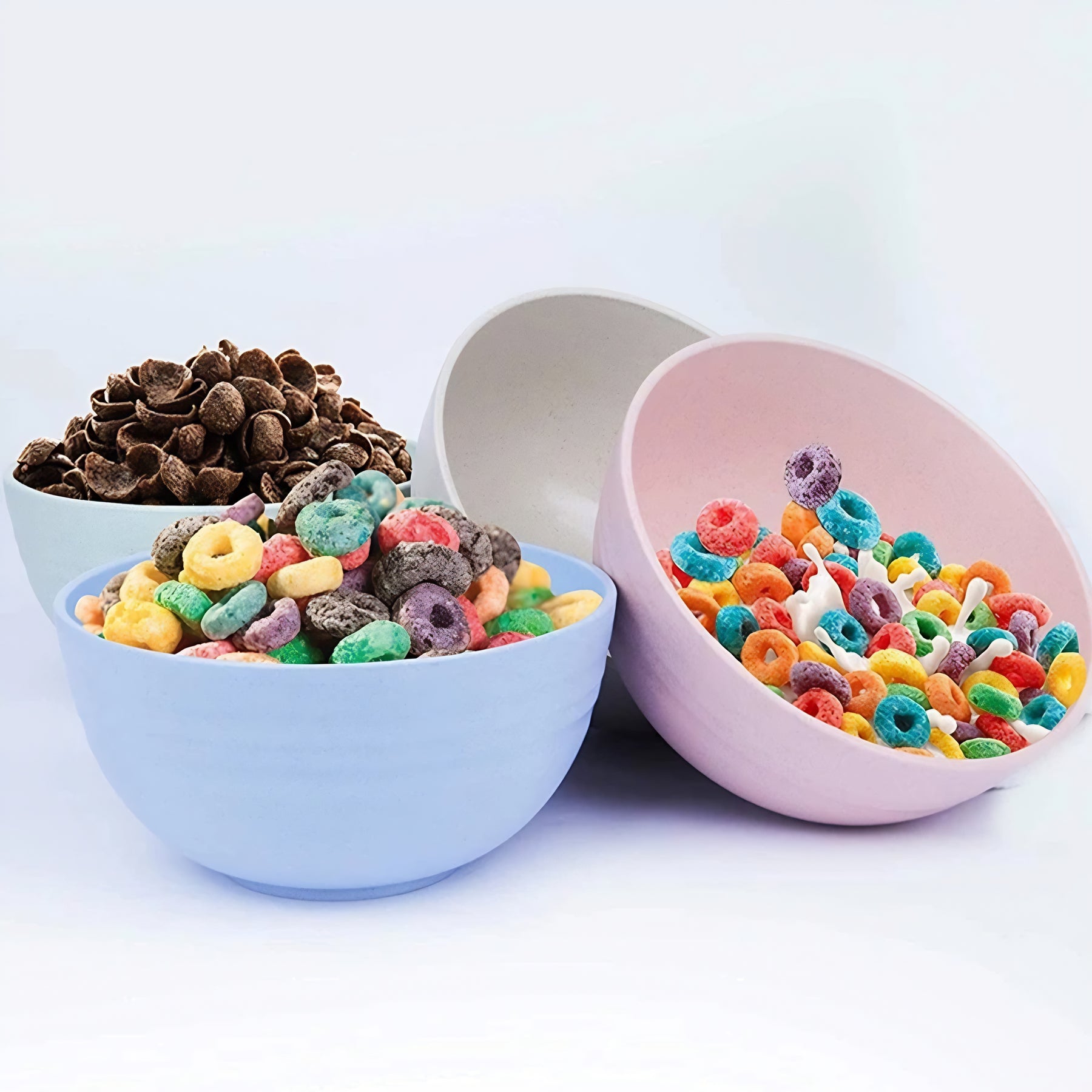Três bowls coloridos em rosa, azul e bege, com dois deles cheios de cereais matinais coloridos.