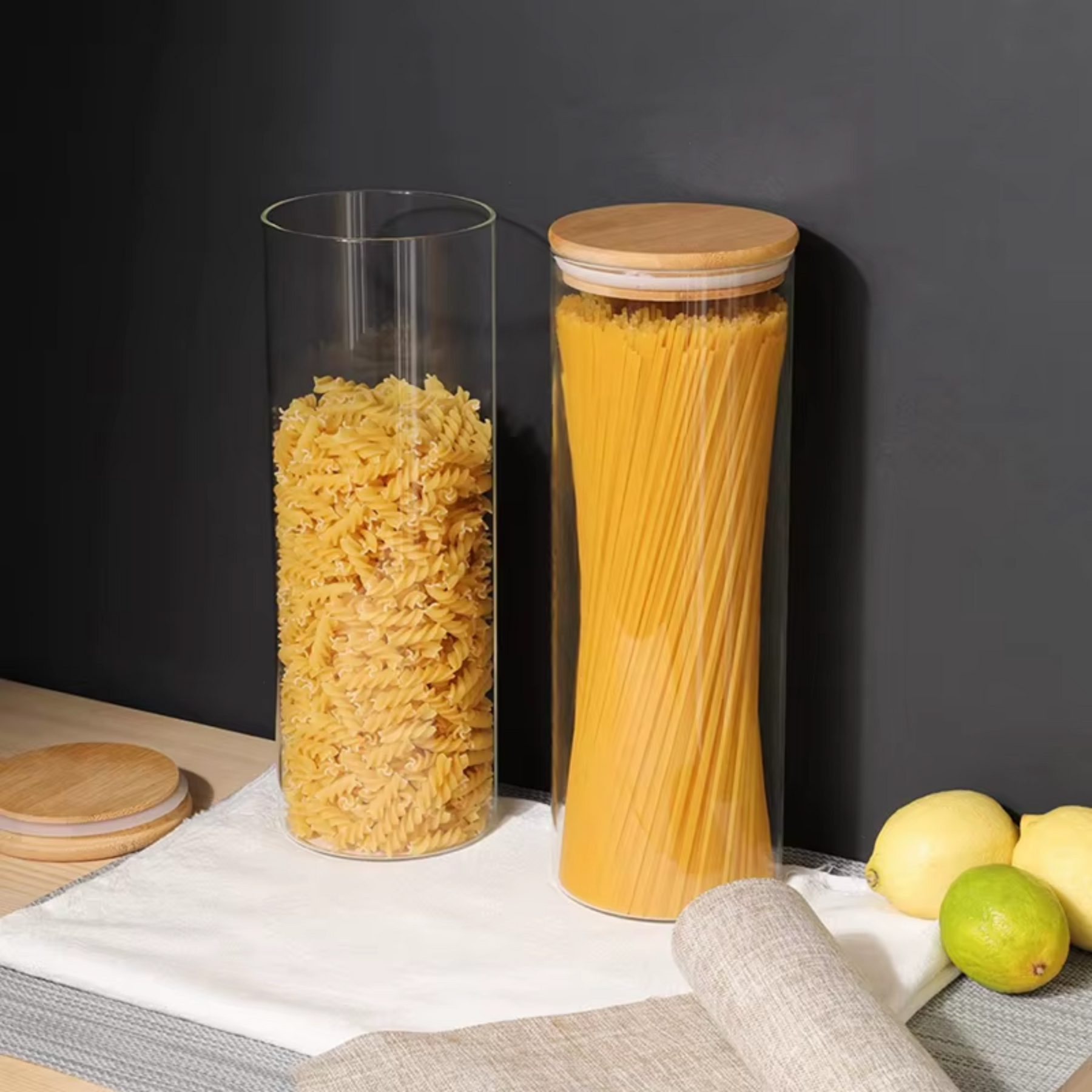 Pote de vidro com tampa de bambu armazenando macarrão espaguete e penne sobre bancada com fundo escuro