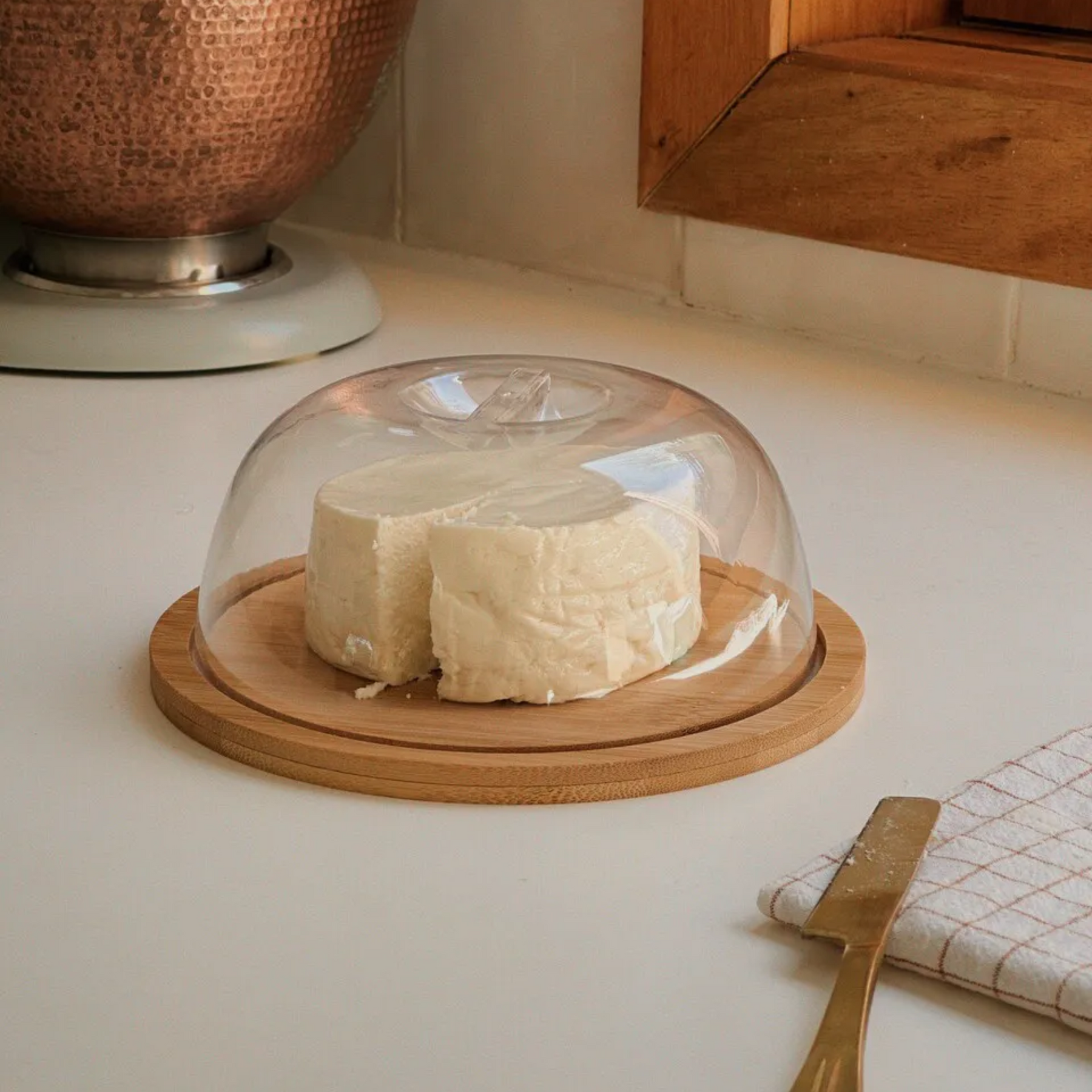 Queijeira de bambu com tampa e queijo branco, disposta em uma bancada de cozinha para armazenamento.