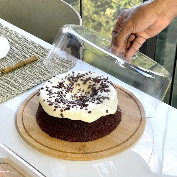 Mão colocando a tampa de acrílico sobre um bolo de chocolate decorado na base da boleira de bambu.