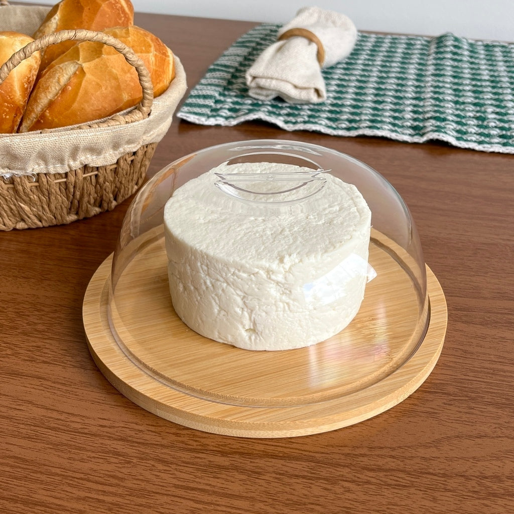 Porta-queijo de bambu redondo com tampa de acrílico sobre mesa de madeira com toalha xadrez ao fundo.