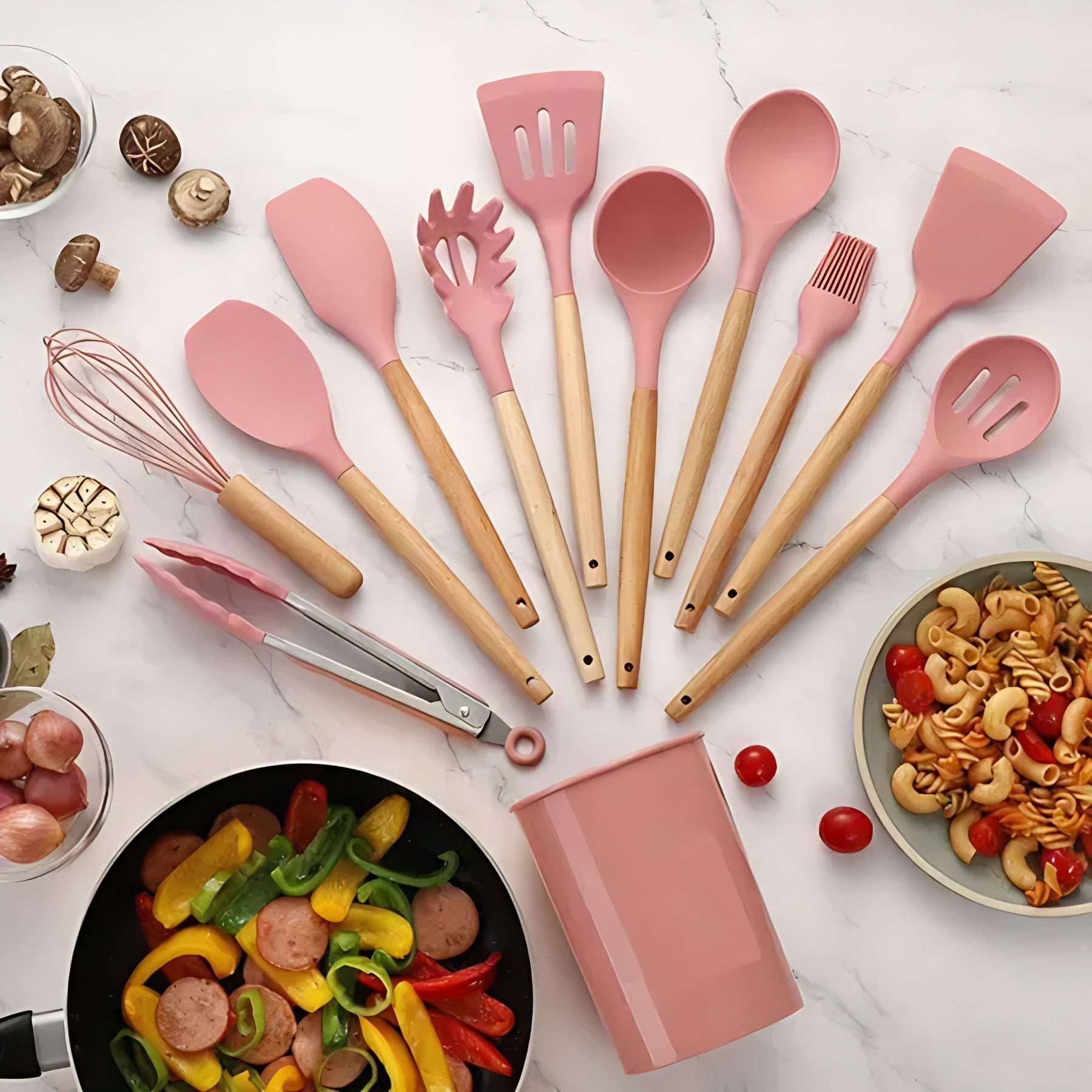 Kit completo de utensílios de cozinha com pontas de silicone rosa e cabos de bambu, ao lado de um pote com macarrão.