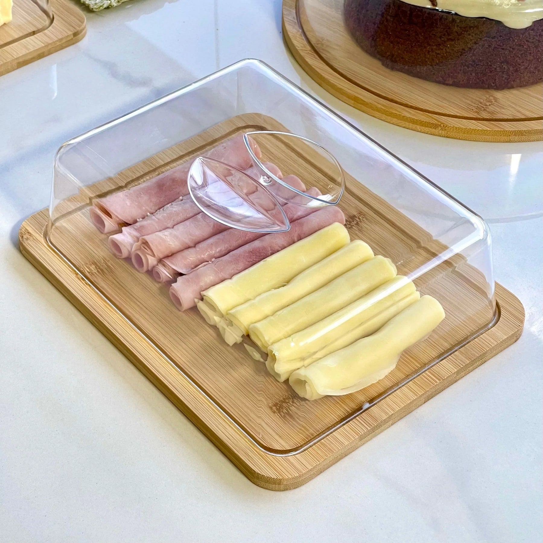 Tábua de frios retangular de bambu com tampa, servindo presunto e queijo em uma mesa de jantar.