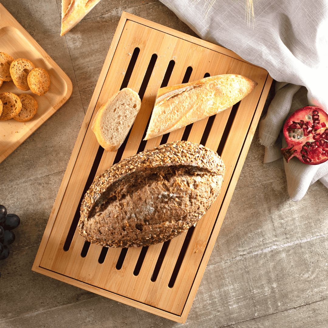 Tábua migalheira de bambu com um pão artesanal grande e fatias de baguete sobre uma toalha de mesa.