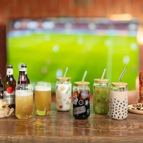 Vários copos de vidro com tampa de bambu enfileirados, servindo drinks em um evento com campo de futebol ao fundo.