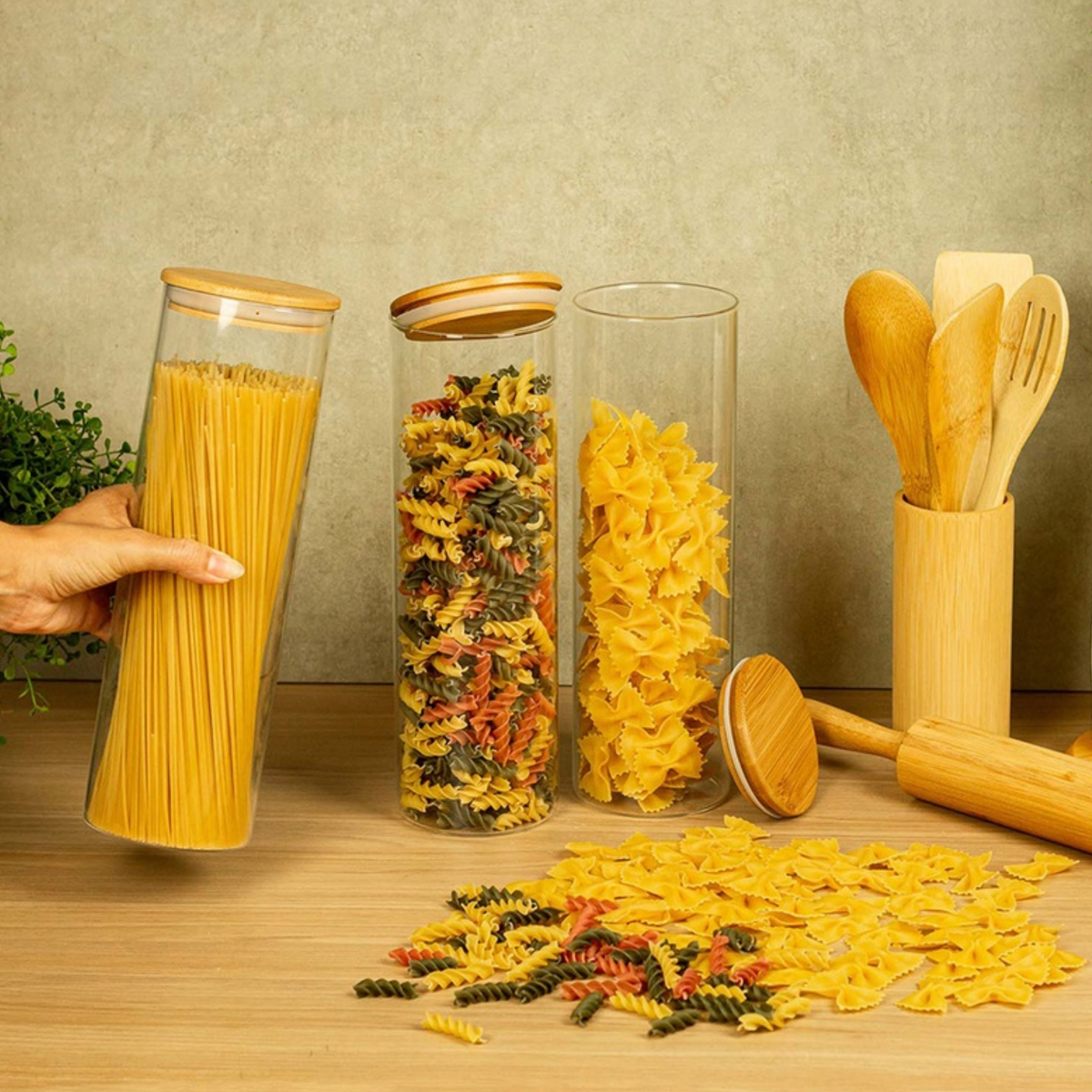 Potes de vidro com tampa de bambu armazenando macarrão espaguete, parafuso colorido e gravatinha sobre bancada

