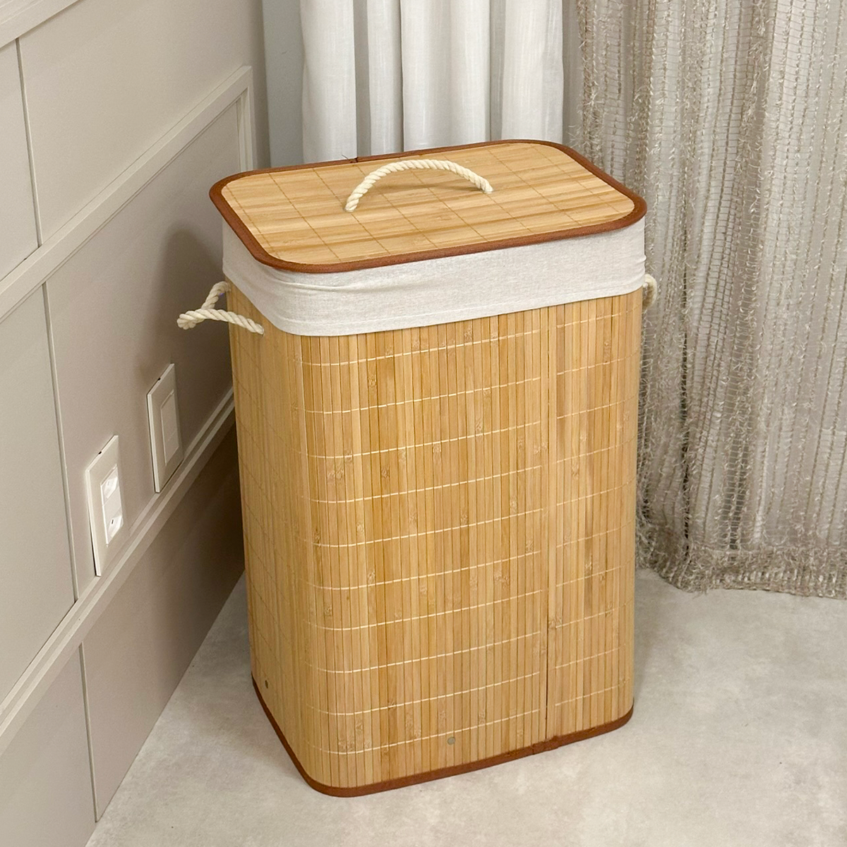Cesto de roupa de bambu com tampa, posicionado no canto de um quarto com parede branca e rodapé.