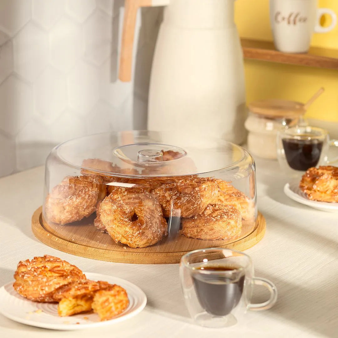 Boleira redonda de bambu com tampa de acrílico, servindo rosquinhas e pães doces em uma mesa de café da manhã.