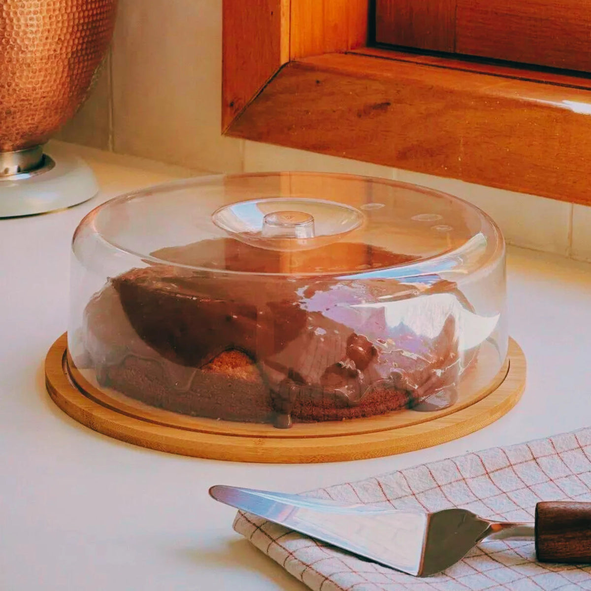 Boleira de bambu com tampa de acrílico armazenando um bolo caseiro em uma bancada de cozinha.