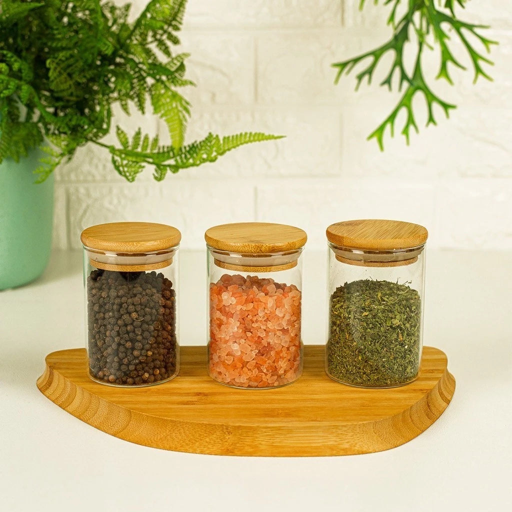 Potes de vidro com tampa de bambu organizando pimenta, sal rosa e ervas, com bandeja de madeira decorativa