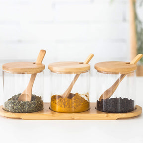 Conjunto de 3 porta-temperos de vidro com tampa e colher de bambu, sobre um suporte, com diferentes especiarias dentro.